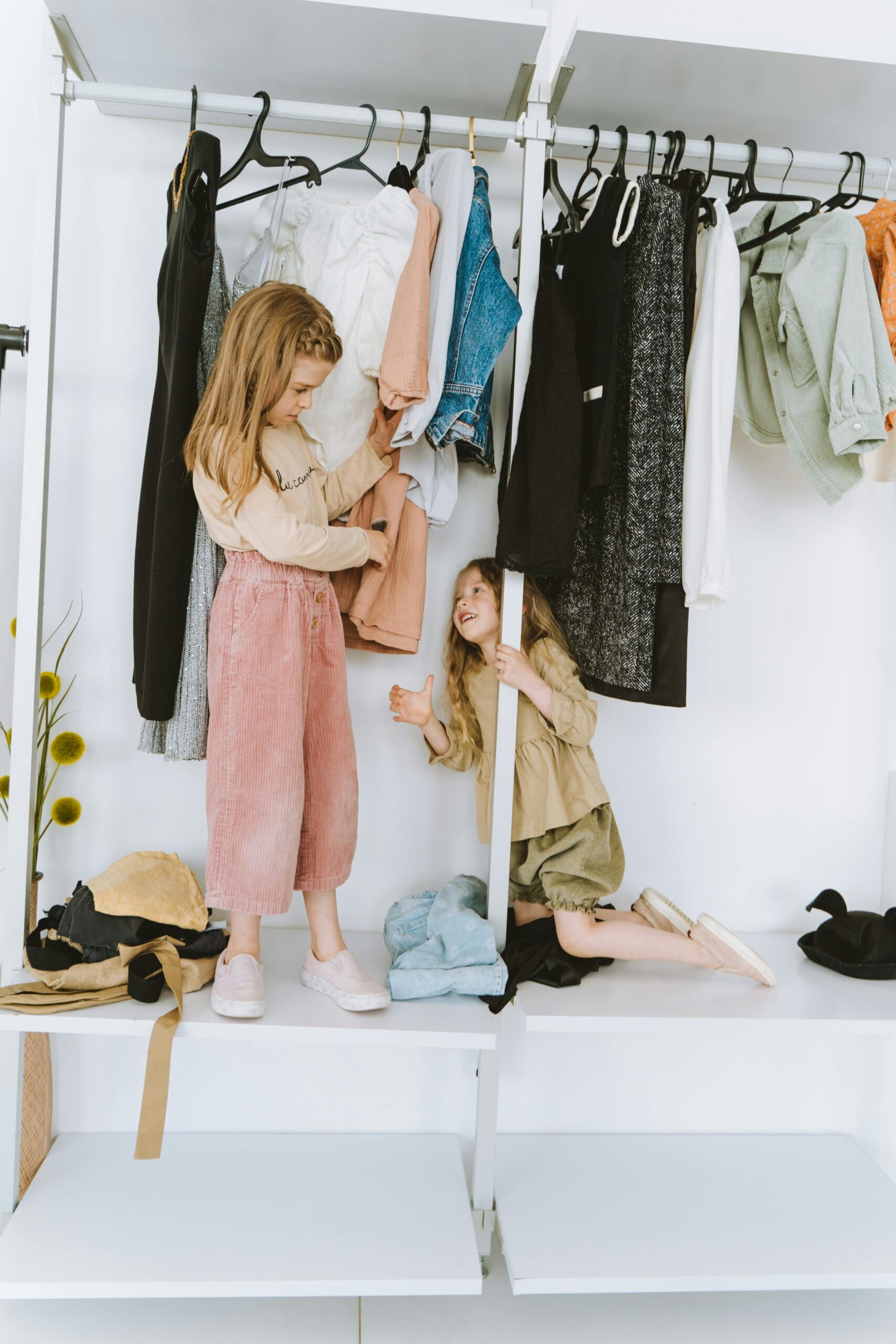 Two smiling girls playing in a wardrobe full of colorful clothes, having fun indoors.